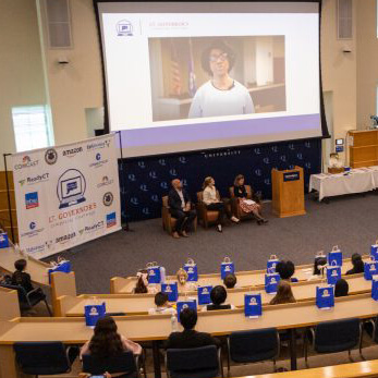 A large screen projects at the front of a Quinnipiac auditorium while people listen seated around