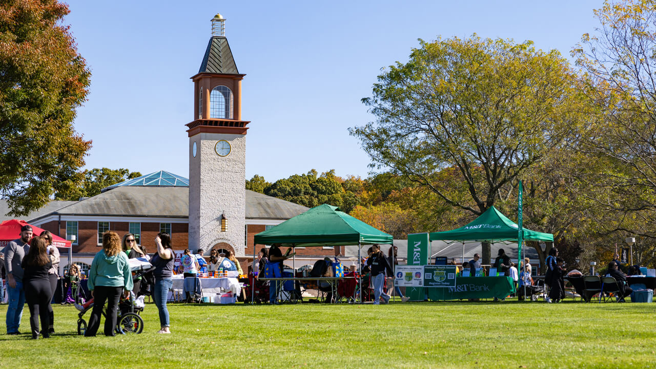 Dozens of people interact on the quad set up with business sun tents with the clocktower in the background